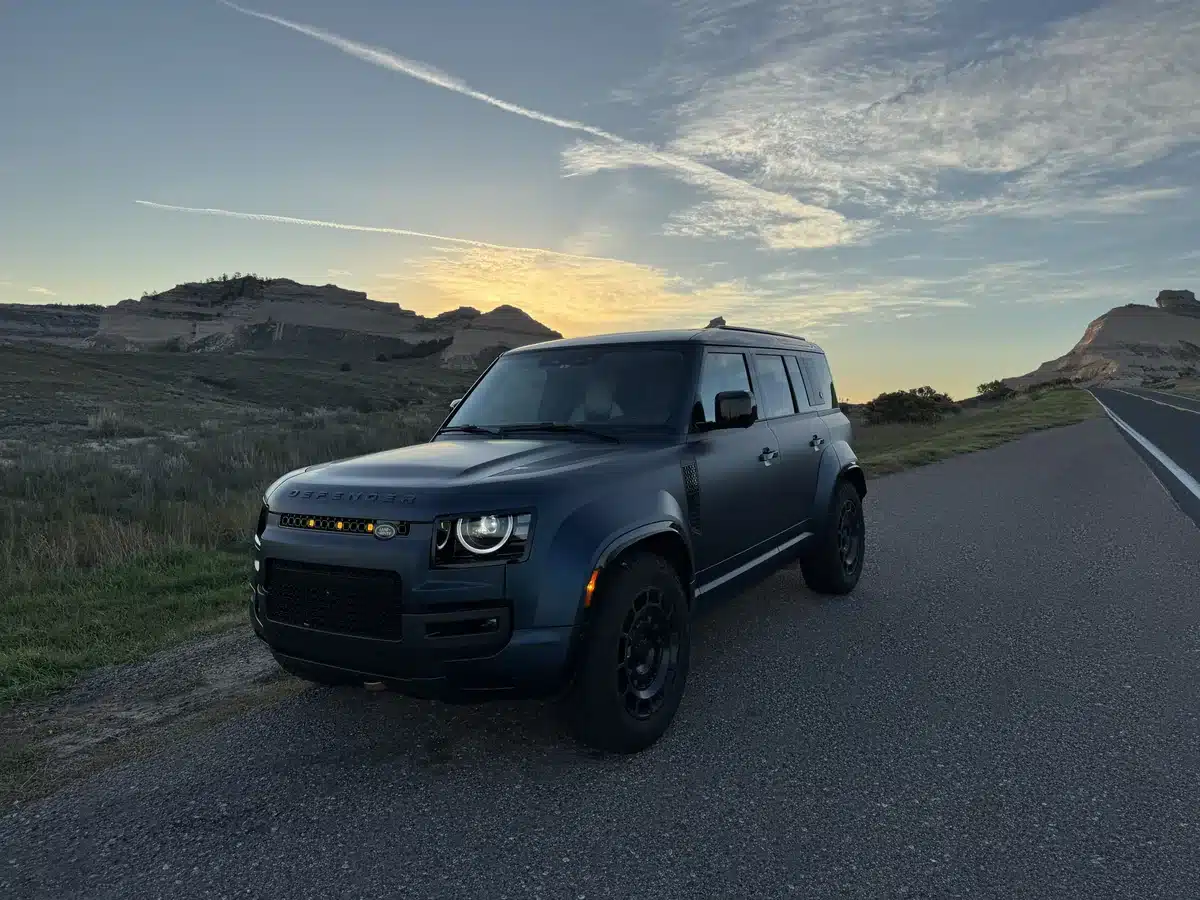 Blue Defender OCTA parked beside a rural road in a front three-quarter view at sunrise.