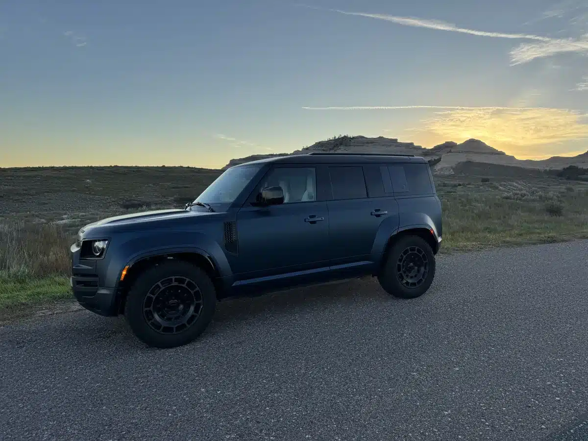 Blue Defender OCTA shown from the driver side beside a rural road at sunrise.